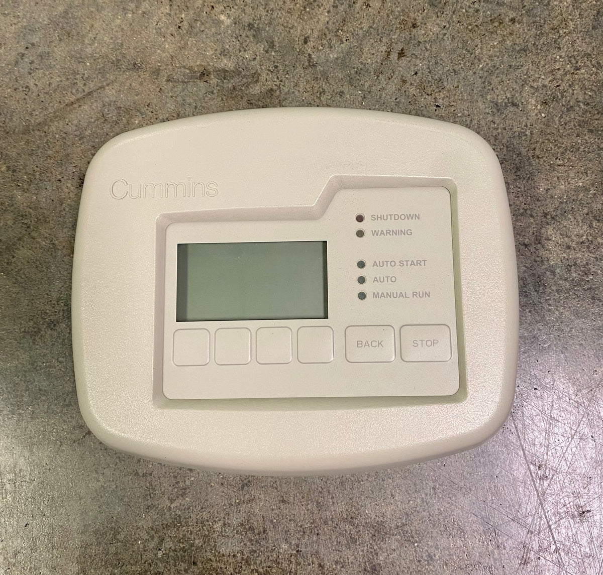 A Cummins engine control module or thermostat cover in a light beige plastic casing, featuring a small digital display screen and buttons, placed on a textured gray concrete surface.