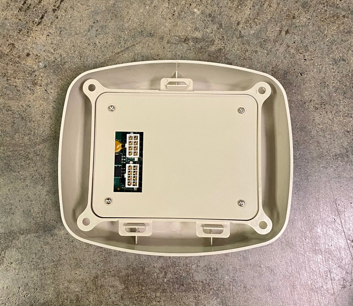 An off-white plastic junction box cover with a small green circuit board connector visible in the center, photographed from above on a gray concrete floor.