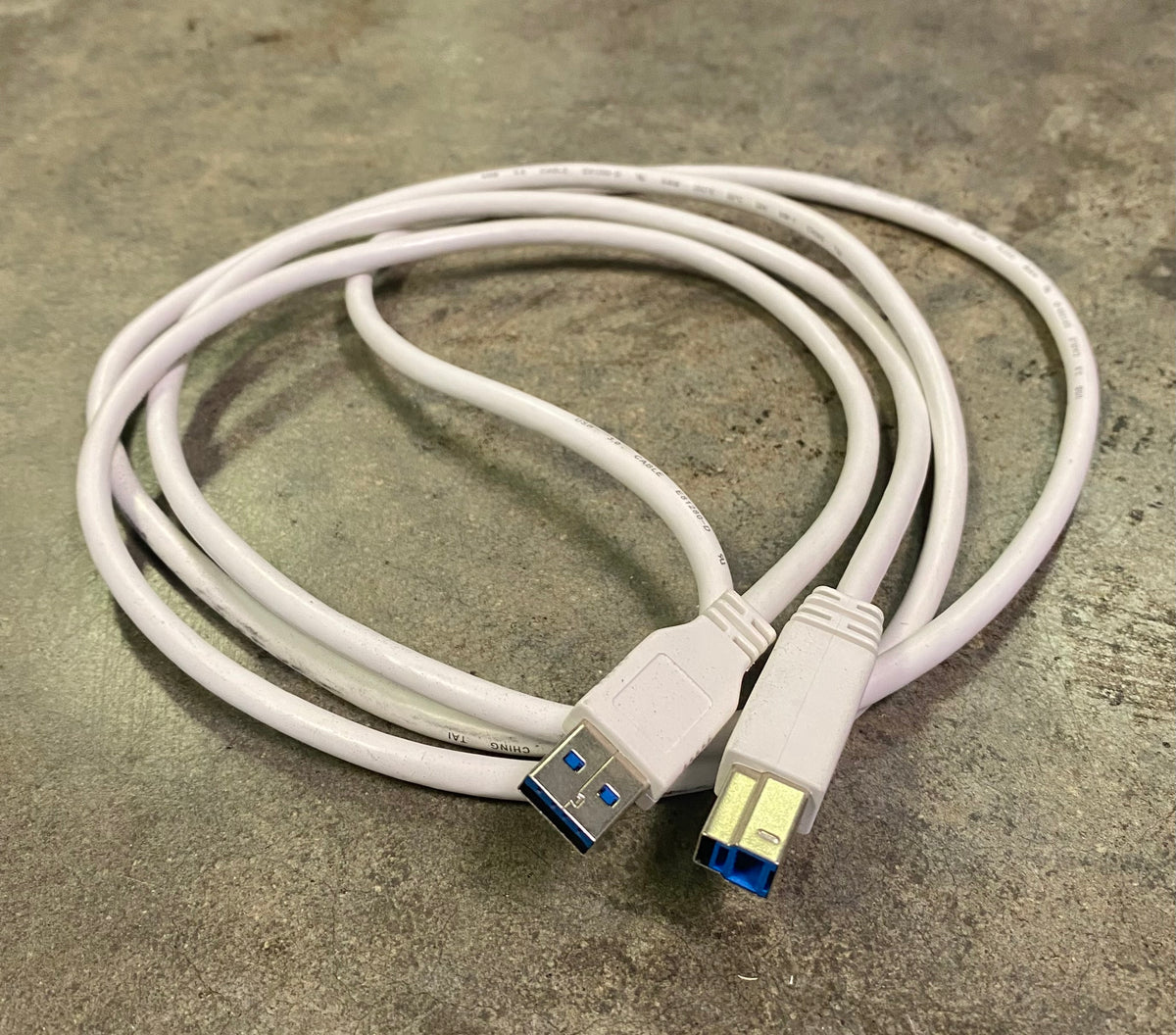 A white USB 3.0 extension cable with two USB-A connectors, one male and one female, coiled on a textured gray concrete surface.