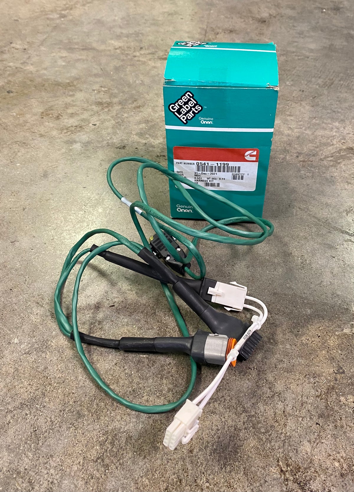 A green and black automotive sensor with attached green and white wires, placed in front of its teal and white branded box on a concrete floor.