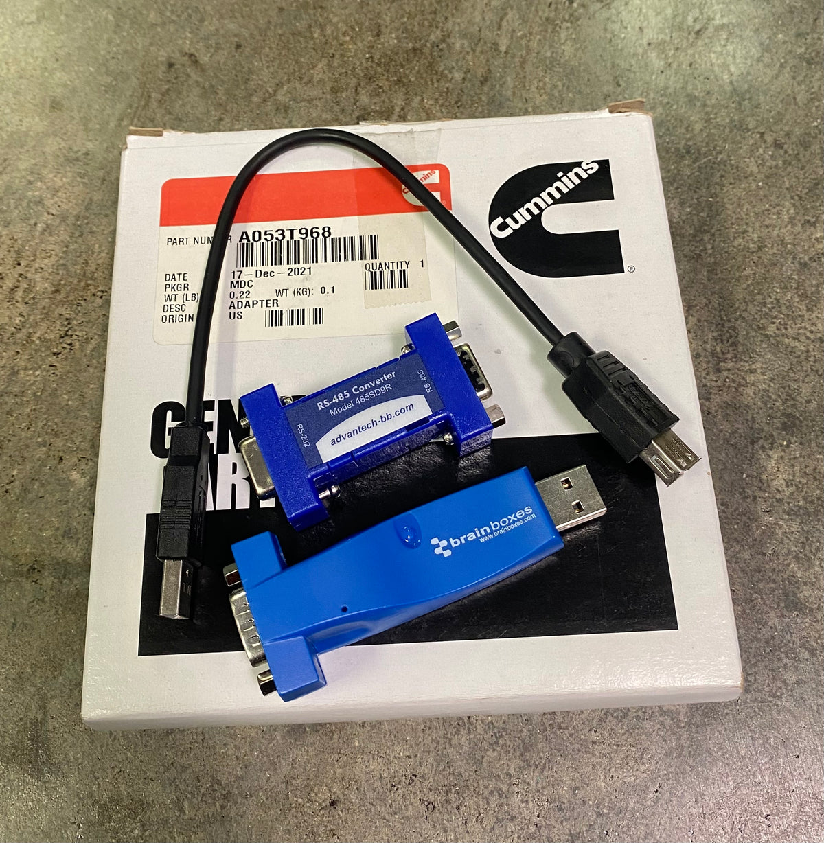 Cummins E-Load Controller adapter cables and two blue brainhouse smart plugs on top of their white packaging box, placed on a gray concrete floor.